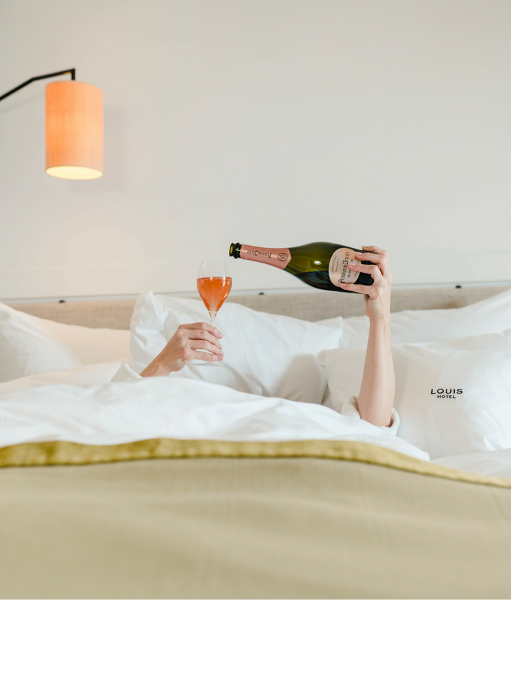 Person im Bett gießt Sekt in ein Glas, umgeben von weißen Kissen im LOUIS Hotel München.
