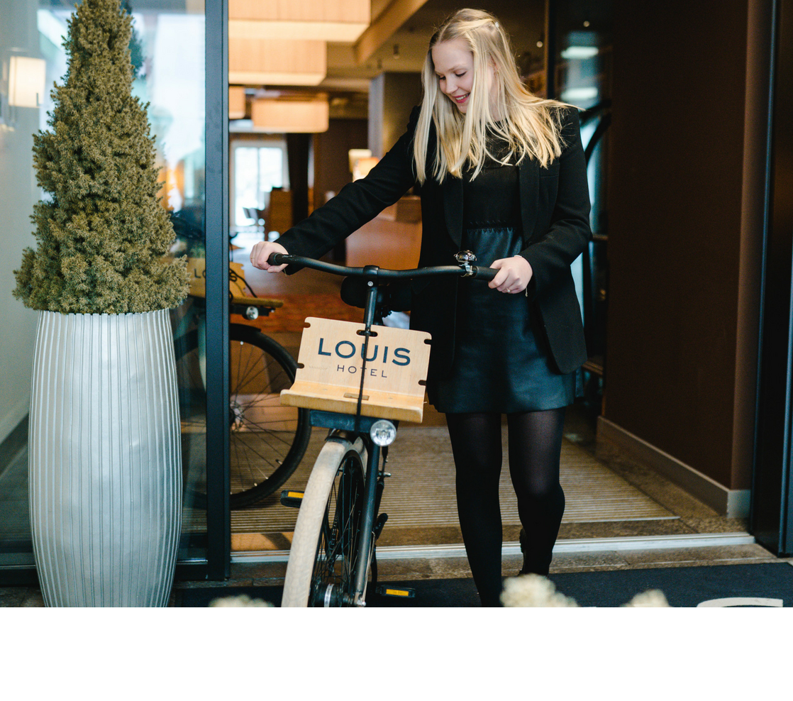 Frau mit Fahrrad am Eingang des LOUIS Hotels München, neben großem Pflanzkübel.