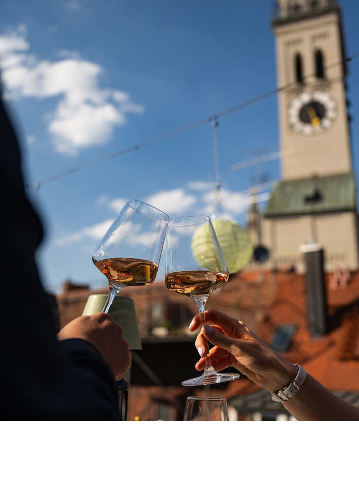 Zwei Menschen stoßen mit Wein auf der Dachterrasse an, im Hintergrund die Sankt Peter Kirche.